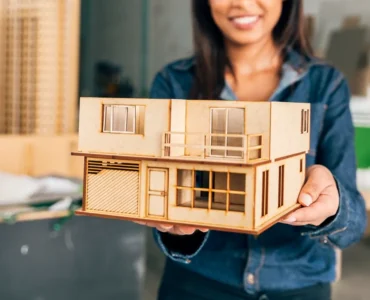 Mulher sorridente segura uma maquete arquitetônica de casa moderna de dois andares feita em madeira, com janelas, varanda e garagem em miniatura.