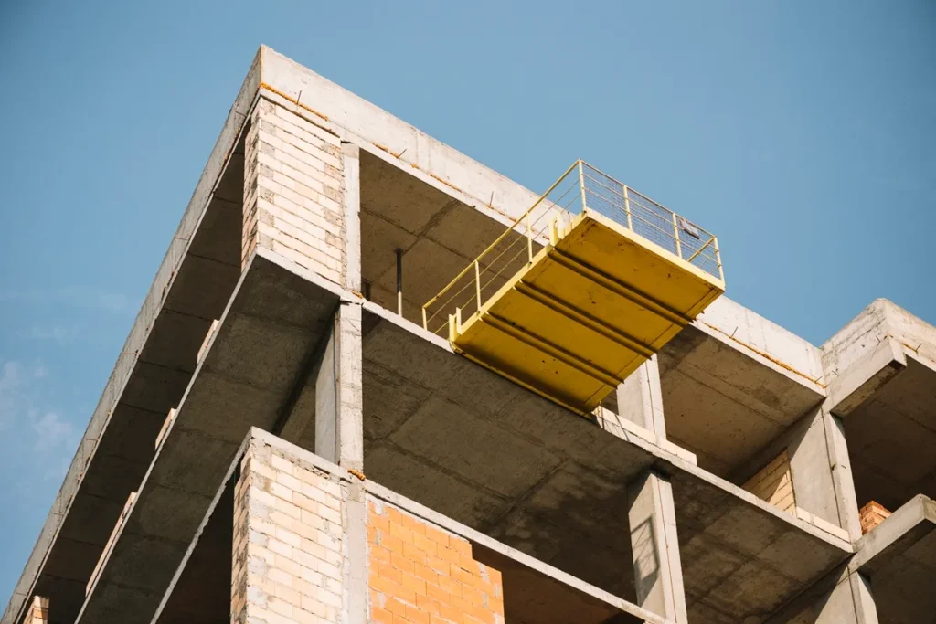 Edifício em construção com estrutura de concreto aparente, paredes de blocos de tijolos e plataforma suspensa amarela projetada para fora da fachada.