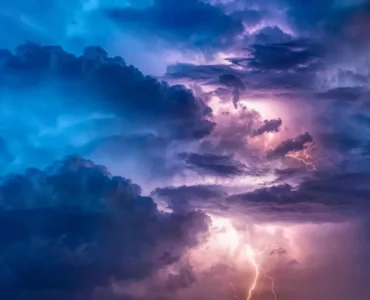 Nuvens densas iluminadas por relâmpagos durante uma tempestade elétrica, com tons de azul e roxo no céu, representando fenômenos meteorológicos intensos.