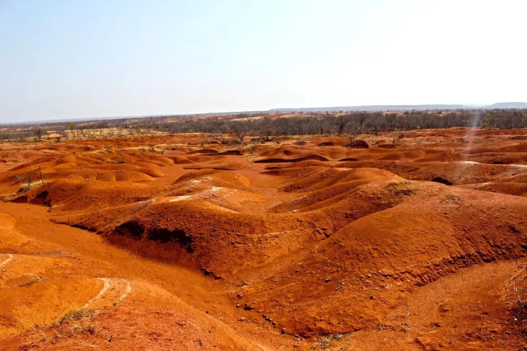 Paisagem com solo avermelhado e ondulado, marcada pela erosão e desertificação em região semiárida, com vegetação escassa ao fundo.