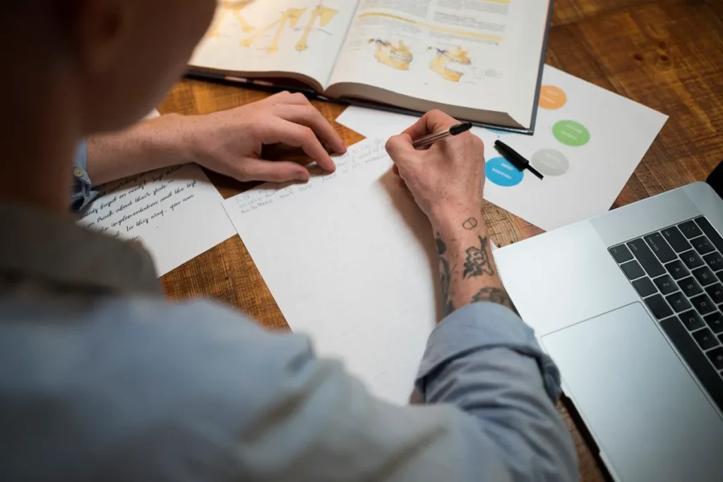Pessoa escrevendo à mão sobre mesa de madeira com livro aberto, papéis e laptop.