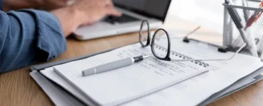 Pessoa digitando no laptop com caderno, óculos e caneta sobre a mesa em ambiente de home office.