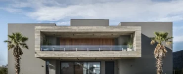 Fachada de casa moderna em estilo brutalista com estrutura de concreto aparente, linhas retas, grandes aberturas de vidro e paisagismo com palmeiras.