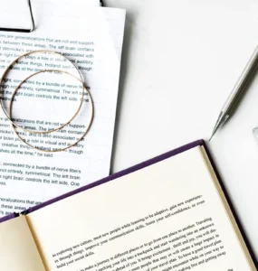 Livro aberto e folhas impressas com textos em inglês sobre a teoria dos hemisférios cerebrais, ao lado de uma caneta prateada, um marcador preto e parte de um teclado de computador sobre uma mesa branca.