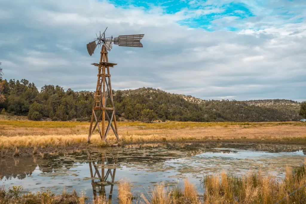Cata-vento de madeira em área rural refletido em uma lagoa rasa, com campos secos e colinas arborizadas ao fundo sob céu parcialmente nublado.