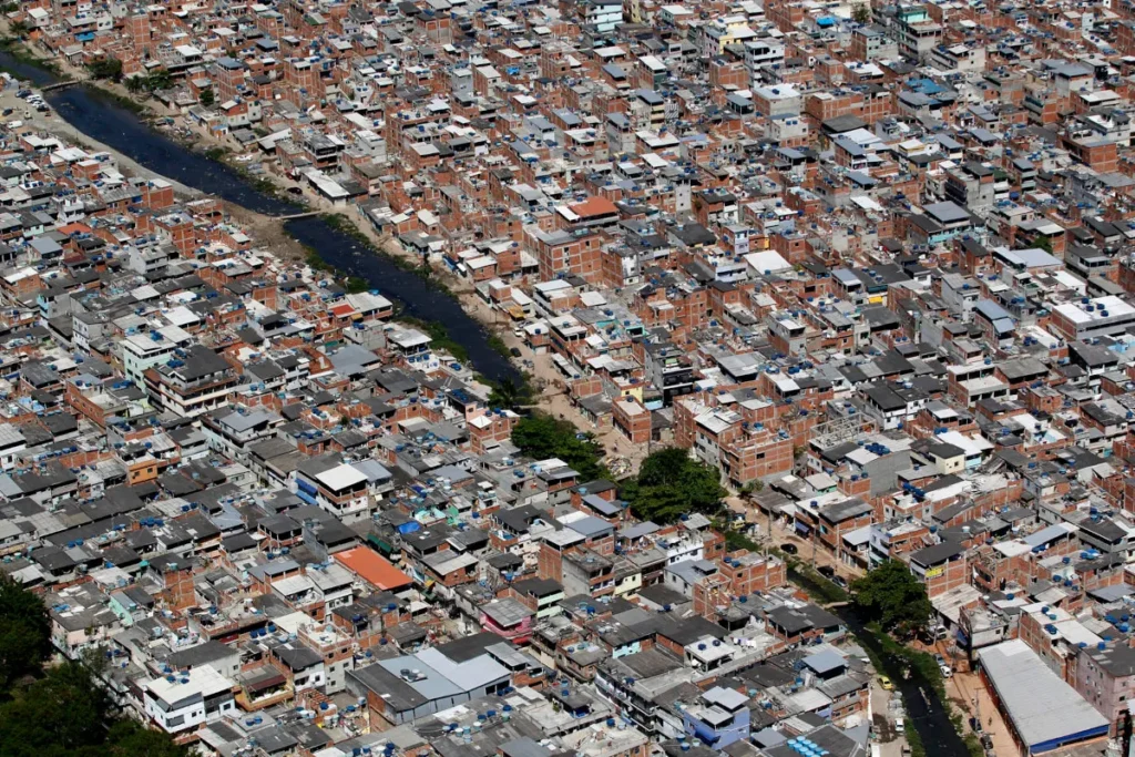 Vista aérea de uma favela brasileira com casas muito próximas, ruas estreitas e um rio que corta a comunidade.