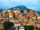 Vista colorida de uma favela brasileira com casas construídas em morros, mostrando edificações irregulares, telhados variados e vegetação ao redor.