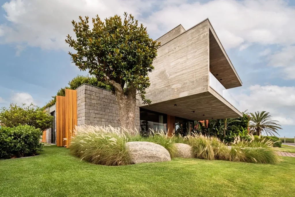 Casa moderna de arquitetura brutalista com estrutura em concreto aparente, detalhes em madeira e jardim com gramado, pedras e vegetação tropical.