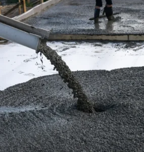 Concreto usinado sendo despejado por calha sobre base de obra, com trabalhador nivelando o material ao fundo.