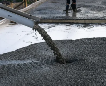 Concreto usinado sendo despejado por calha sobre base de obra, com trabalhador nivelando o material ao fundo.