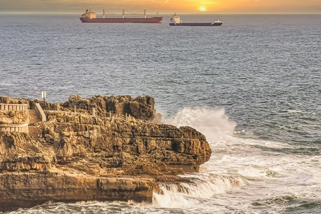 Ondas do mar batendo contra falésias rochosas ao pôr do sol, com navios ao fundo navegando sobre as correntes marítimas.
