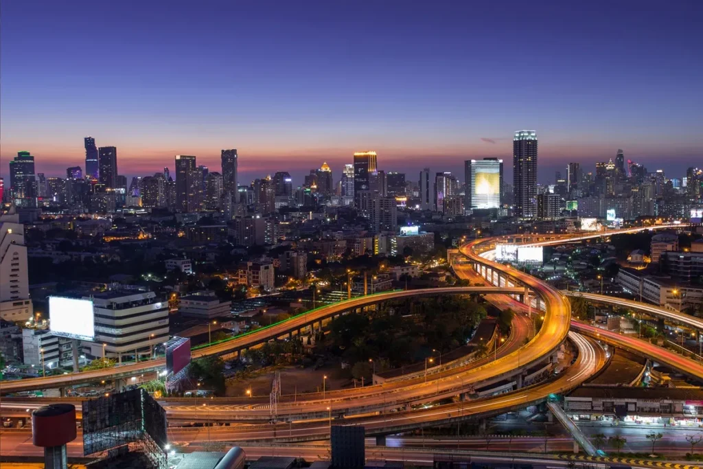 Vista aérea noturna de uma megacidade com arranha-céus iluminados e rodovias curvas com tráfego intenso.