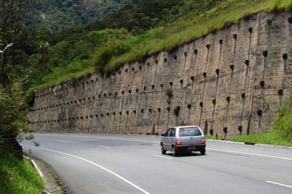 Muro de arrimo de concreto ao lado de uma rodovia em área de serra, com carro passando.