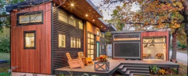 Tiny house moderna com fachada de madeira e metal, deck amplo com poltronas, iluminação embutida e miniestúdio anexo com design que imita um amplificador.
