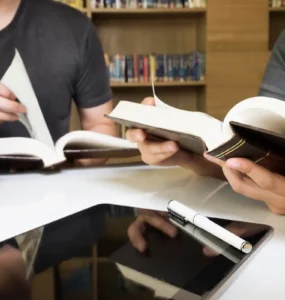 Duas pessoas estudando em uma biblioteca, lendo livros abertos sobre uma mesa com tablet e caneta, representando pesquisa e produção acadêmica.