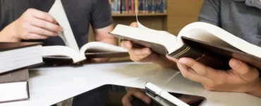Duas pessoas estudando em uma biblioteca, lendo livros abertos sobre uma mesa com tablet e caneta, representando pesquisa e produção acadêmica.