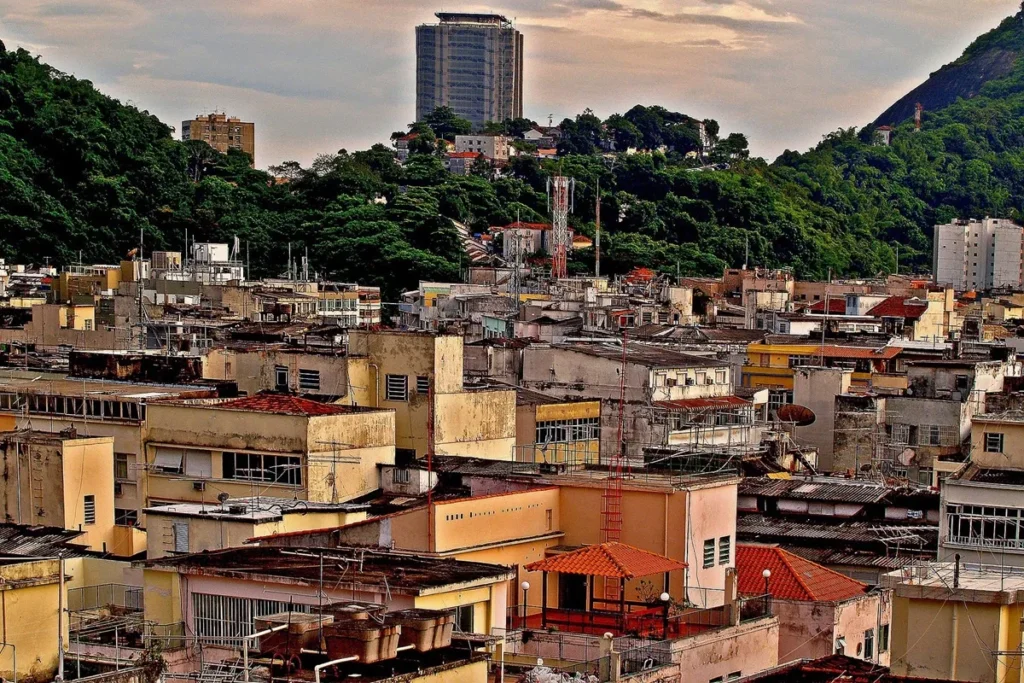 Vista panorâmica de um bairro com moradias densas e irregulares em contraste com áreas verdes e prédios altos ao fundo.