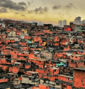 Vista aérea de uma comunidade com casas de tijolo aparente, sob um céu nublado ao entardecer, com prédios distantes ao fundo.