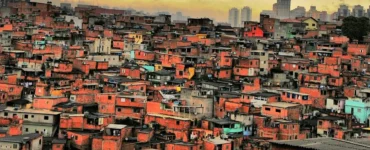 Vista aérea de uma comunidade com casas de tijolo aparente, sob um céu nublado ao entardecer, com prédios distantes ao fundo.