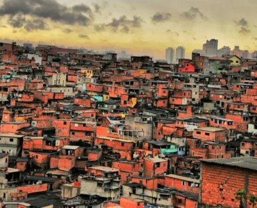 Vista aérea de uma comunidade com casas de tijolo aparente, sob um céu nublado ao entardecer, com prédios distantes ao fundo.