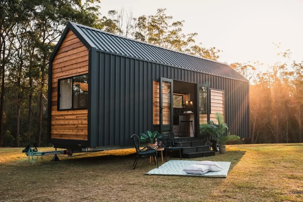 Tiny house moderna com fachada de metal escuro e madeira, portas abertas, área externa com cadeiras e tapete ao ar livre ao pôr do sol.