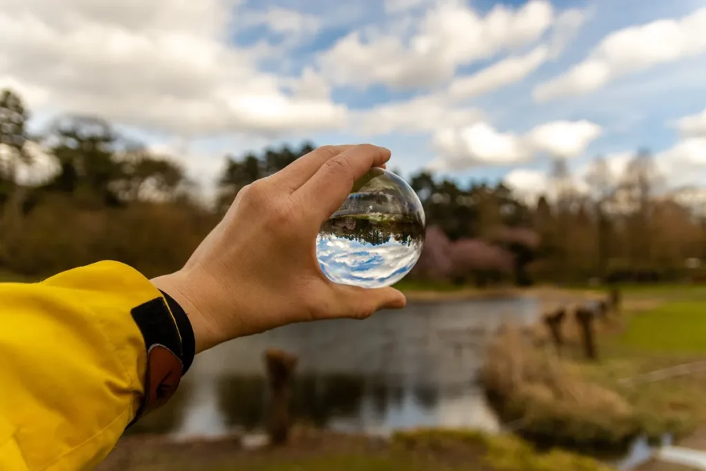 Mão segurando esfera de cristal que reflete paisagem com lago e céu, simbolizando o ciclo da água na natureza
