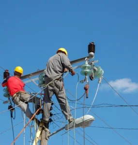 Dois eletricistas com capacete realizando manutenção em poste de energia, manuseando cabos e equipamentos de alta tensão sob céu azul.