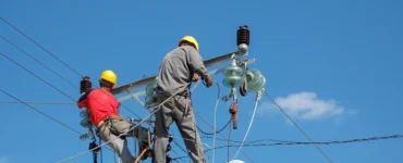 Dois eletricistas com capacete realizando manutenção em poste de energia, manuseando cabos e equipamentos de alta tensão sob céu azul.