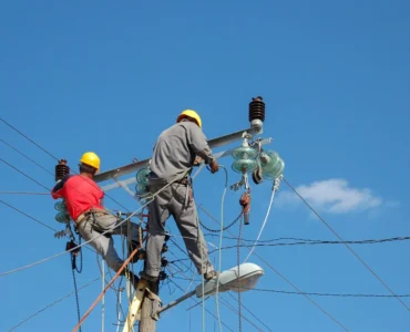 Dois eletricistas com capacete realizando manutenção em poste de energia, manuseando cabos e equipamentos de alta tensão sob céu azul.