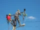 Dois eletricistas com capacete realizando manutenção em poste de energia, manuseando cabos e equipamentos de alta tensão sob céu azul.