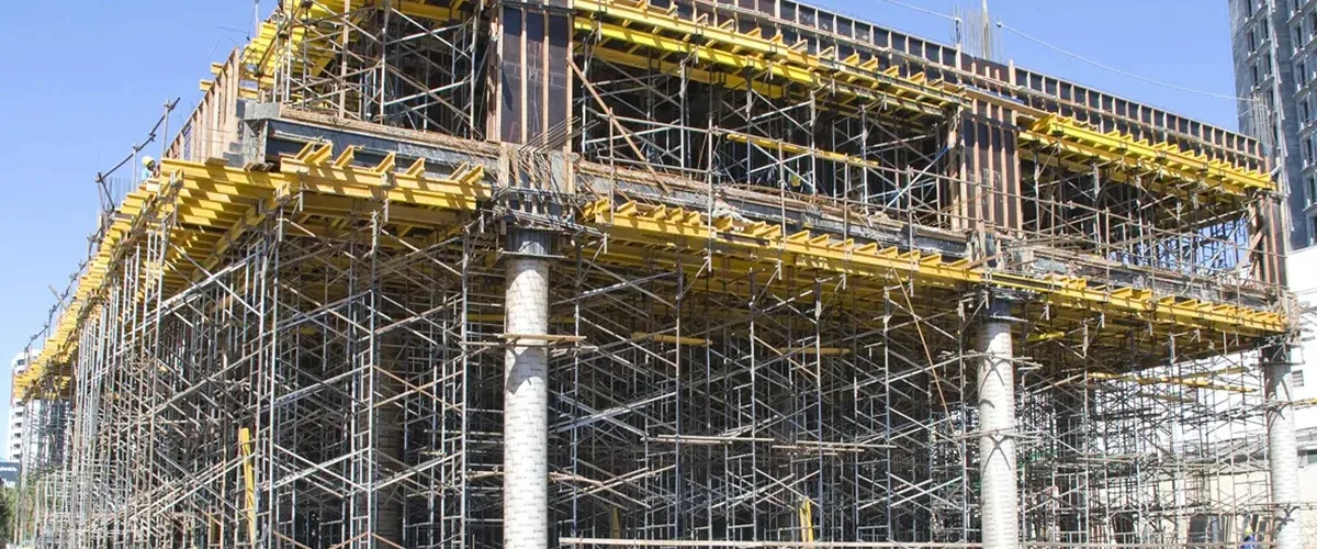 Estrutura de escoramento metálico sustentando laje em construção de prédio, com andaimes, vigas de madeira e trabalhadores realizando montagem.
