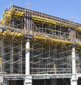 Estrutura de escoramento metálico sustentando laje em construção de prédio, com andaimes, vigas de madeira e trabalhadores realizando montagem.