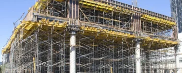 Estrutura de escoramento metálico sustentando laje em construção de prédio, com andaimes, vigas de madeira e trabalhadores realizando montagem.