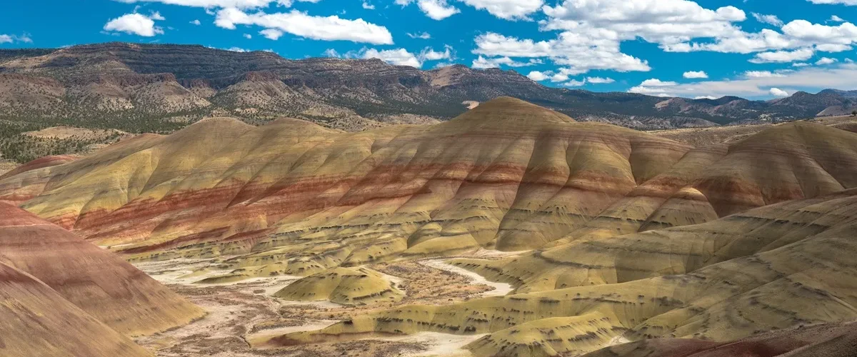 Formações geológicas coloridas com camadas de solo em tons de amarelo, vermelho e marrom em uma paisagem árida com colinas onduladas sob céu azul com nuvens brancas.