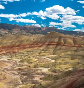 Formações geológicas coloridas com camadas de solo em tons de amarelo, vermelho e marrom em uma paisagem árida com colinas onduladas sob céu azul com nuvens brancas.