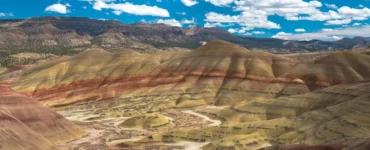 Formações geológicas coloridas com camadas de solo em tons de amarelo, vermelho e marrom em uma paisagem árida com colinas onduladas sob céu azul com nuvens brancas.