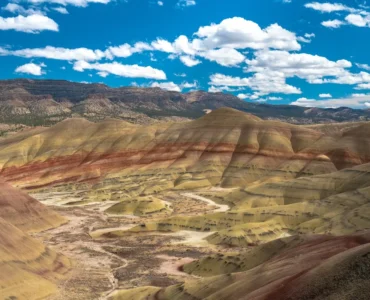 Formações geológicas coloridas com camadas de solo em tons de amarelo, vermelho e marrom em uma paisagem árida com colinas onduladas sob céu azul com nuvens brancas.