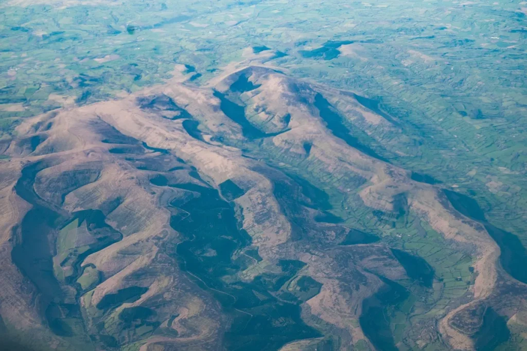 Vista aérea de relevo montanhoso com encostas suaves, áreas agrícolas em tons de verde e vales profundos moldados pela erosão natural.