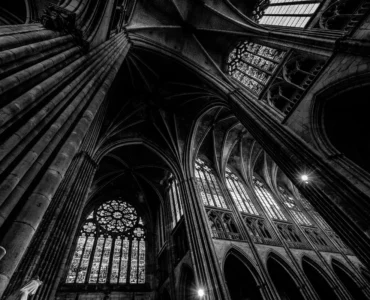 Interior de catedral gótica com colunas altas, arcos ogivais e vitrais iluminados, fotografado em preto e branco.