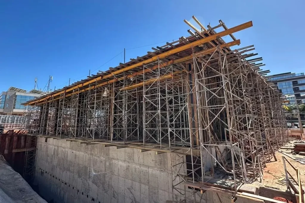 Estrutura de escoramento metálico sustentando laje de concreto em construção, com vigas de madeira e andaimes montados em vários níveis.