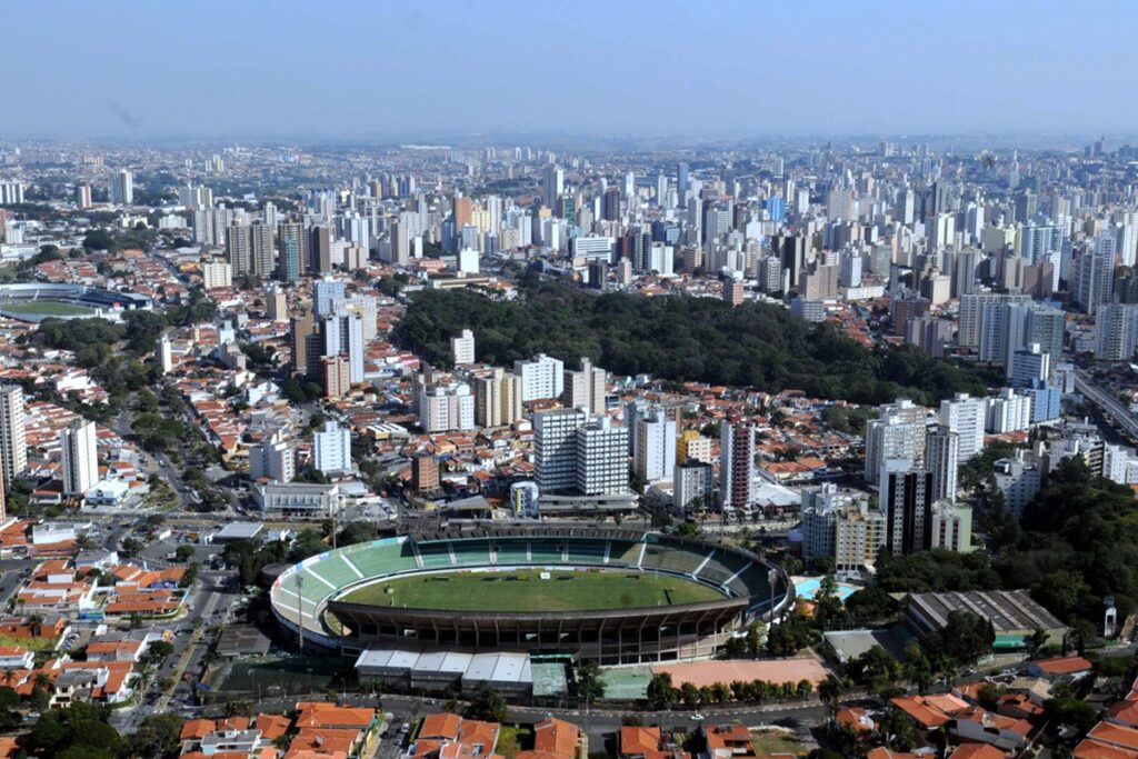 Vista aérea de uma cidade brasileira com estádio de futebol ao centro, bairros residenciais e muitos prédios ao fundo.