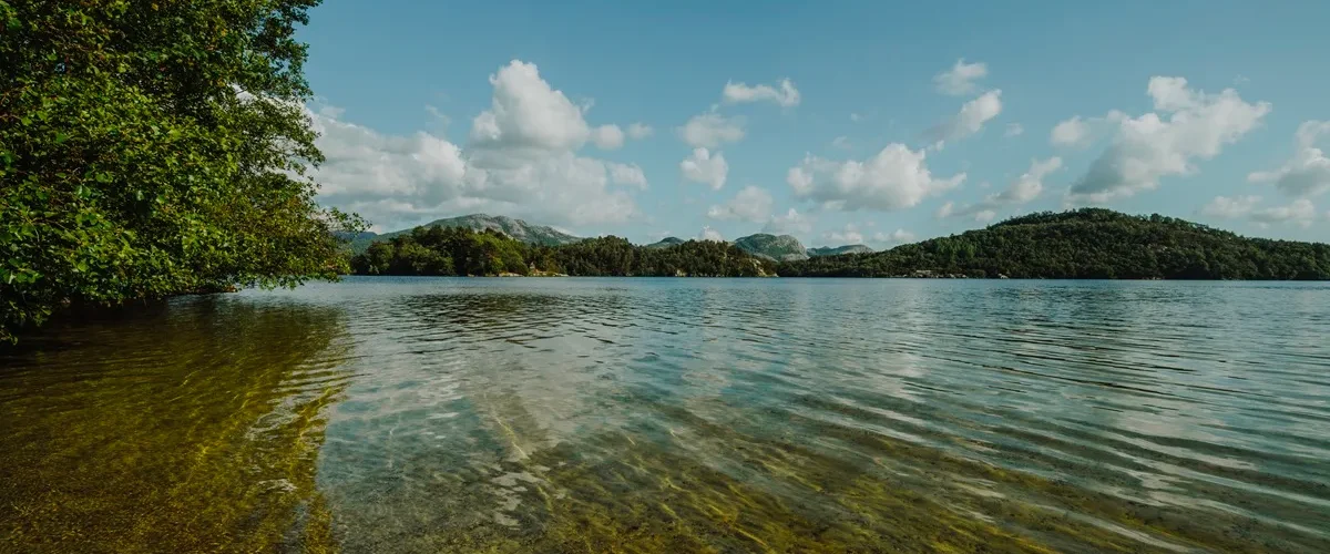 Lago de água cristalina com vegetação lateral, montanhas ao fundo e céu azul com nuvens brancas.