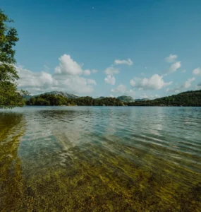 Lago de água cristalina com vegetação lateral, montanhas ao fundo e céu azul com nuvens brancas.