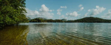 Lago de água cristalina com vegetação lateral, montanhas ao fundo e céu azul com nuvens brancas.