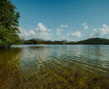 Lago de água cristalina com vegetação lateral, montanhas ao fundo e céu azul com nuvens brancas.