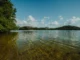 Lago de água cristalina com vegetação lateral, montanhas ao fundo e céu azul com nuvens brancas.