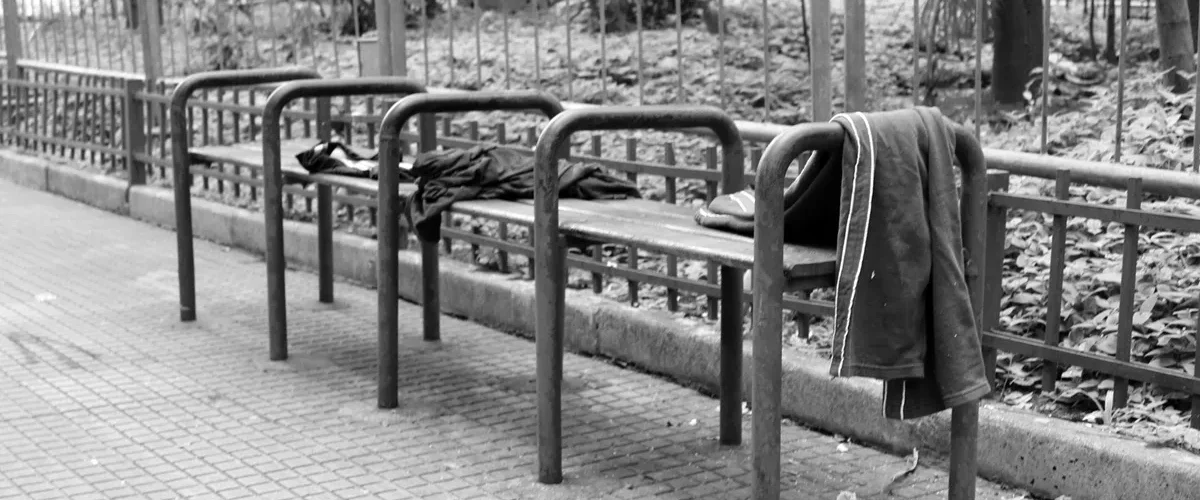 Banco de praça com barras metálicas divisórias impedindo que pessoas se deitem, com roupas deixadas sobre o assento.