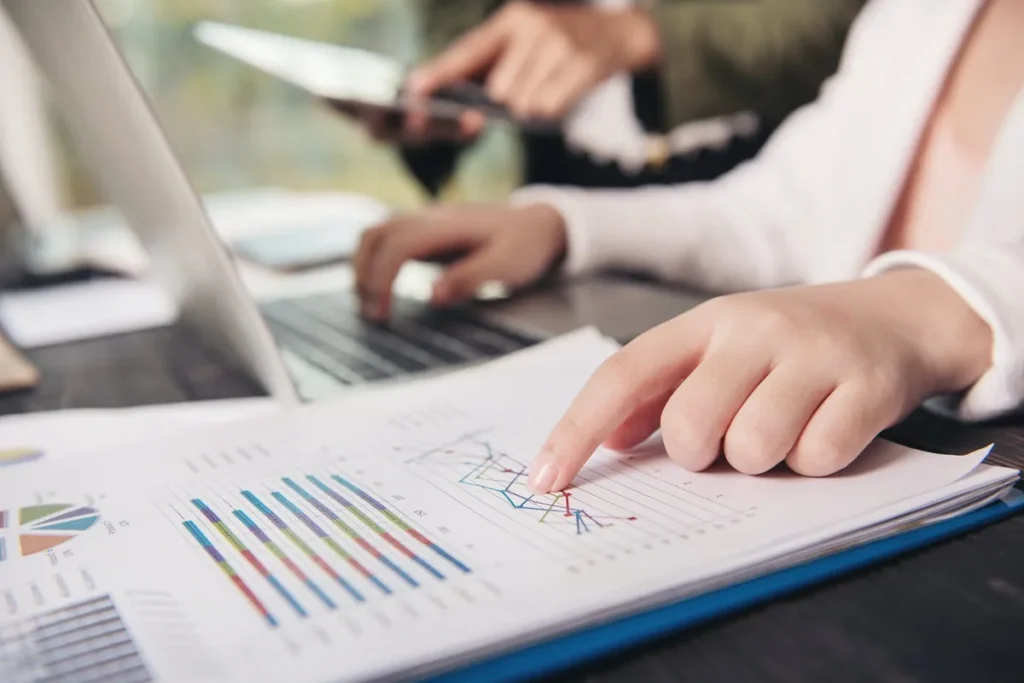 Mão apontando para gráficos coloridos em relatórios financeiros enquanto outra pessoa trabalha no notebook durante reunião.