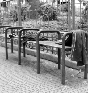 Banco de praça com barras metálicas divisórias impedindo que pessoas se deitem, com roupas deixadas sobre o assento.