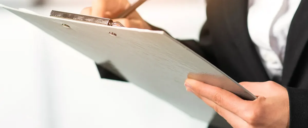 Pessoa com traje social escrevendo em uma prancheta, destacando mãos, caneta e documento em ambiente corporativo.